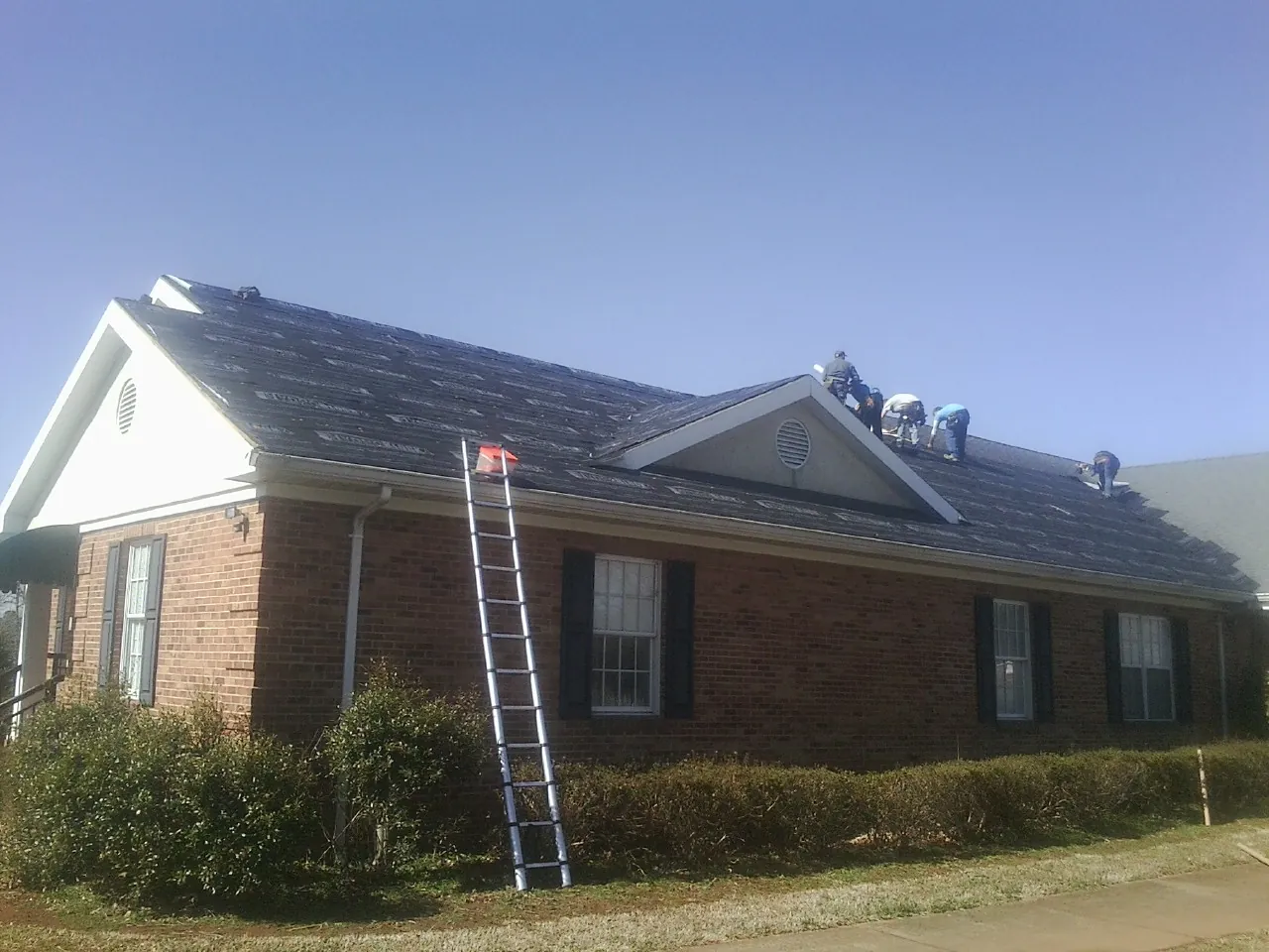Crew tearing shingles off a brick ranch roof with a ladder set — Emergency Roof Repair work by our crew in Six Lakes