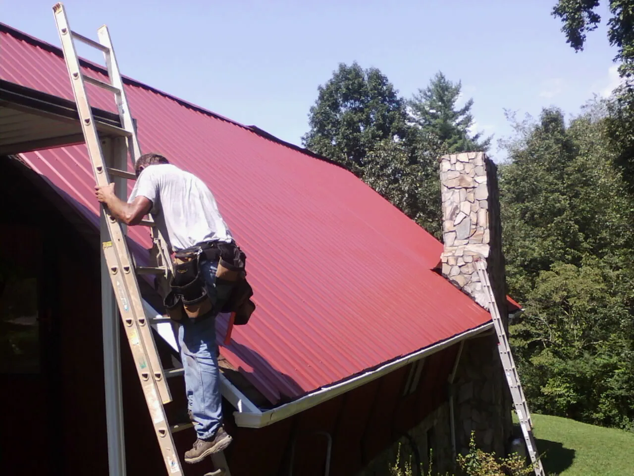 Roofer on a ladder beside a red metal roof and stone chimney — Soffit & Fascia Repair by Roofing Brothers in Six Lakes